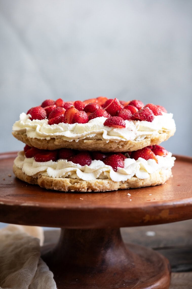 A double-layered strawberry shortcake cake with whipped cream and fresh strawberries sits on a wooden cake stand against a light, blurred background.