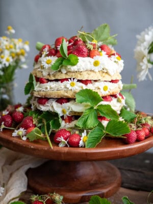 A strawberry shortcake cake decorated with whipped cream, strawberries, green leaves, and small white flowers sits on a wooden cake stand, surrounded by more strawberries and foliage.