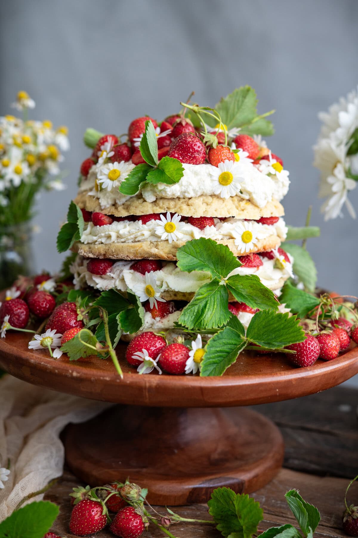 A strawberry shortcake cake decorated with whipped cream, strawberries, green leaves, and small white flowers sits on a wooden cake stand, surrounded by more strawberries and foliage.