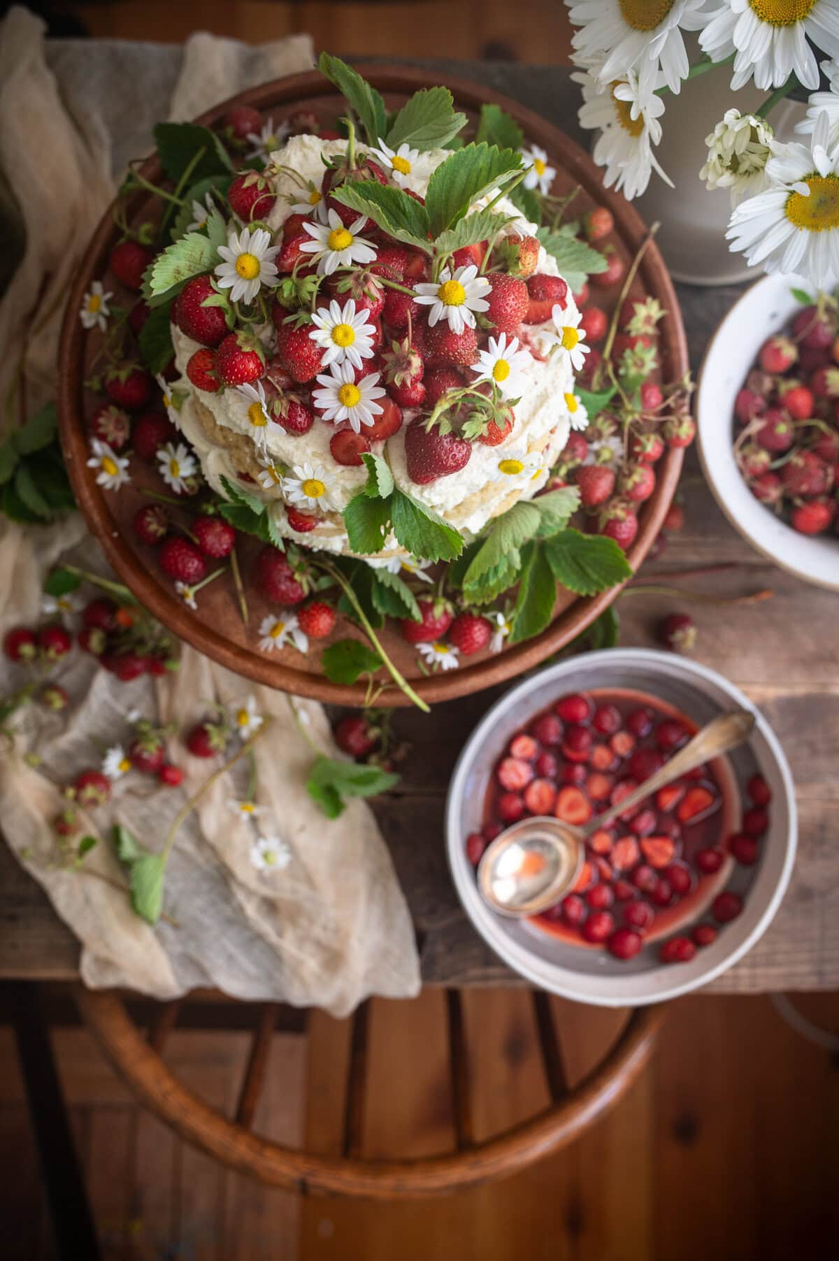 A rustic table features a strawberry shortcake cake topped with whipped cream, fresh strawberries, and daisies, surrounded by more strawberries, green leaves, a white bowl with strawberries and syrup, and a vase of daisies.