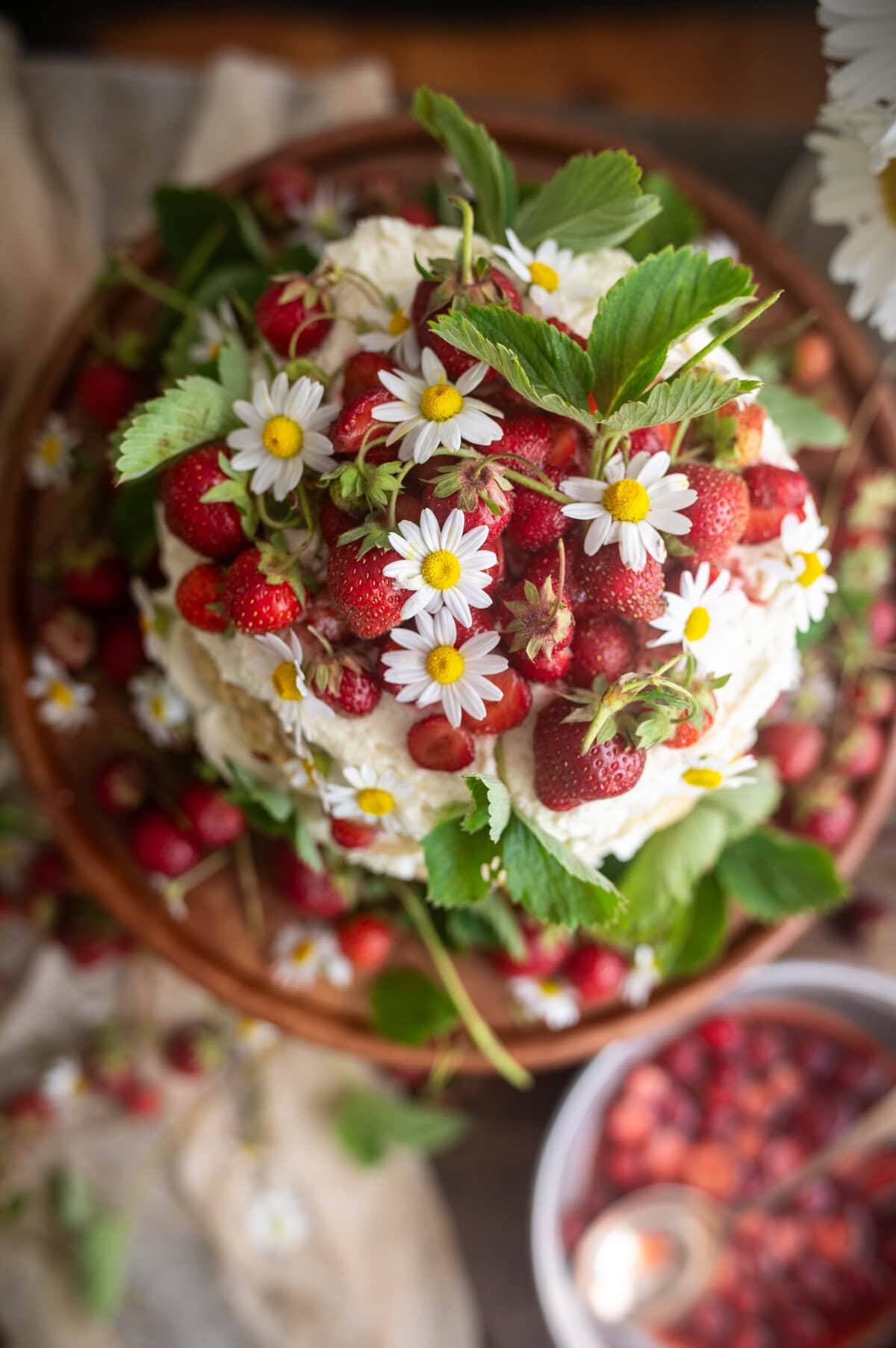 A round strawberry shortcake cake topped with fresh strawberries, whipped cream, and small white daisies sits on a wooden plate, with scattered strawberries and flowers around it.