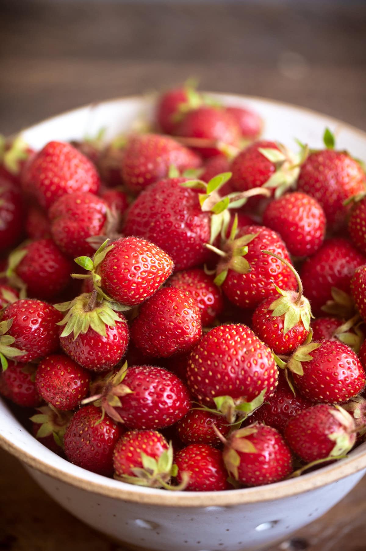 A white ceramic bowl filled with fresh, ripe red strawberries with green stems, perfect for topping a homemade strawberry shortcake cake, sits on a rustic wooden surface.