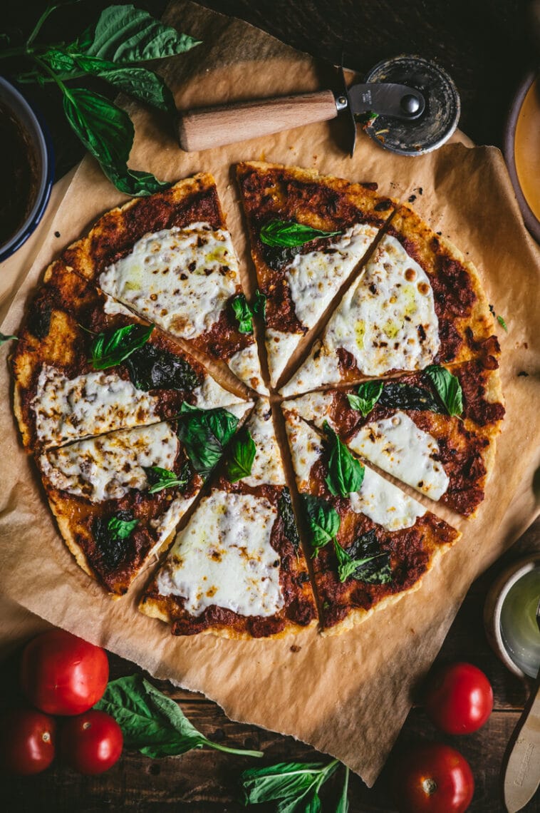 Top down shot of the cooked and sliced gluten-free cassava-chickpea pizza on parchment paper. The pizza is topped with tomato sauce, fresh mozzarella cheese and fresh green basil leaves.