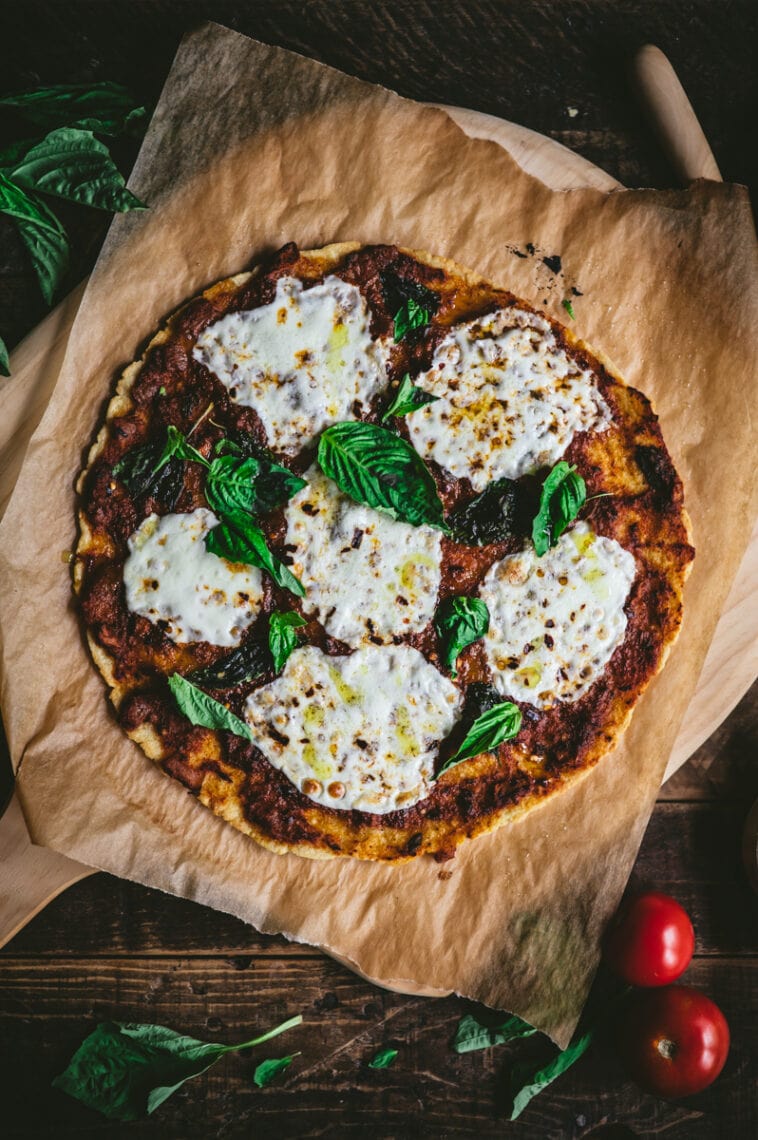 Top down shot of the cooked gluten-free cassava-chickpea pizza on parchment paper. The pizza is topped with tomato sauce, fresh mozzarella cheese and fresh green basil leaves.