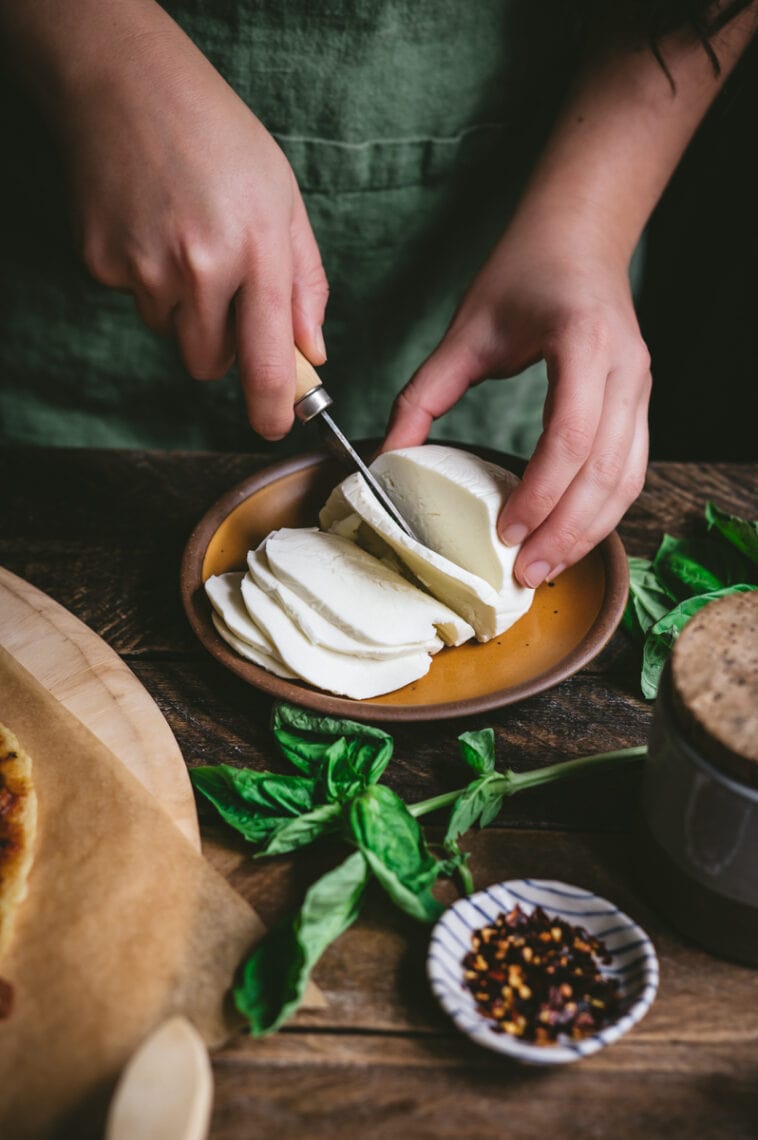Kaity slices into a ball of fresh mozzarella cheese on a small cermaic plate. Fresh basil leaves are strewn about on the surrounding table.