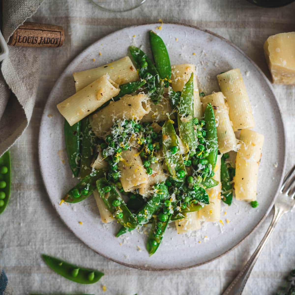 Top down view of a plate of pasta and green peas with lemon and parmesan cheese plated on a white plate that is on a beige linen tablecloth with an antique silver plated fork to the right of the plate. The tabletop is styled with a chunk of parmesan cheese, a glass of white wine, light pink taper candles in brass candlesticks, a small ceramic berry basket filled with fresh snap peas, sprigs of thyme, a whole lemon, and a clear glass vase of green stemmed pink tulip flowers.