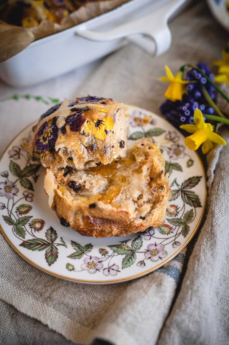 Closeup view of a single hot cross bun spilt open and spread with butter and orange marmalade on a small china plate with a strawberry plant pattern.