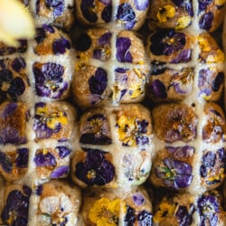 Top down closeup view of baked hot cross buns decorated with fresh edible viola flowers and white flour paste crosses in a pan.