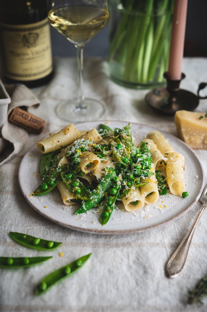 A plate of pasta and green peas with lemon and parmesan cheese plated on a white plate that is on a beige linen tablecloth with an antique silver plated fork to the right of the plate. The tabletop is styled with a chunk of parmesan cheese, a glass of white wine, light pink taper candles in brass candlesticks, a small ceramic berry basket filled with fresh snap peas, sprigs of thyme, and a clear glass vase of green stemmed pink tulip flowers.