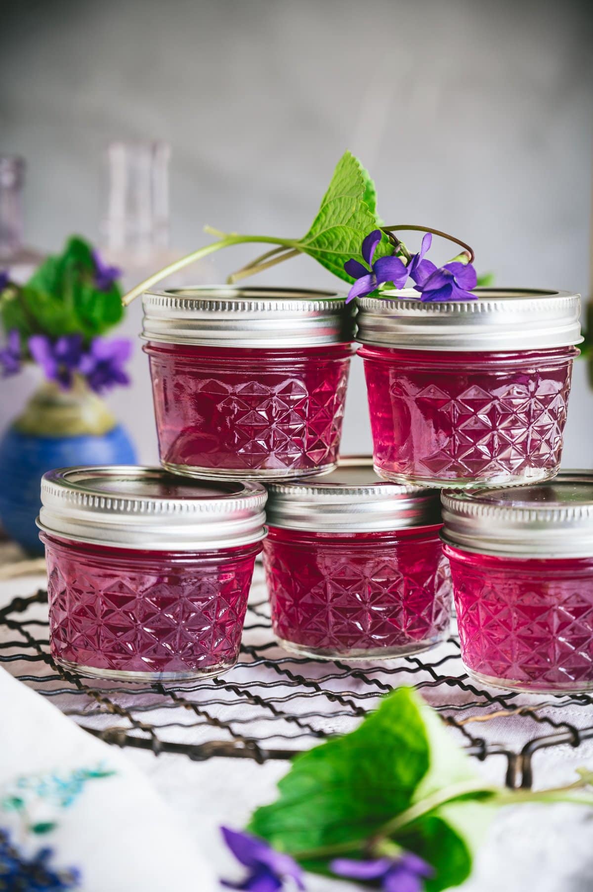 Closeup shot of 5 small quilted mason jars with metal screw top lids filled with purple/pink colored wild violet jelly stacked on top of each other.