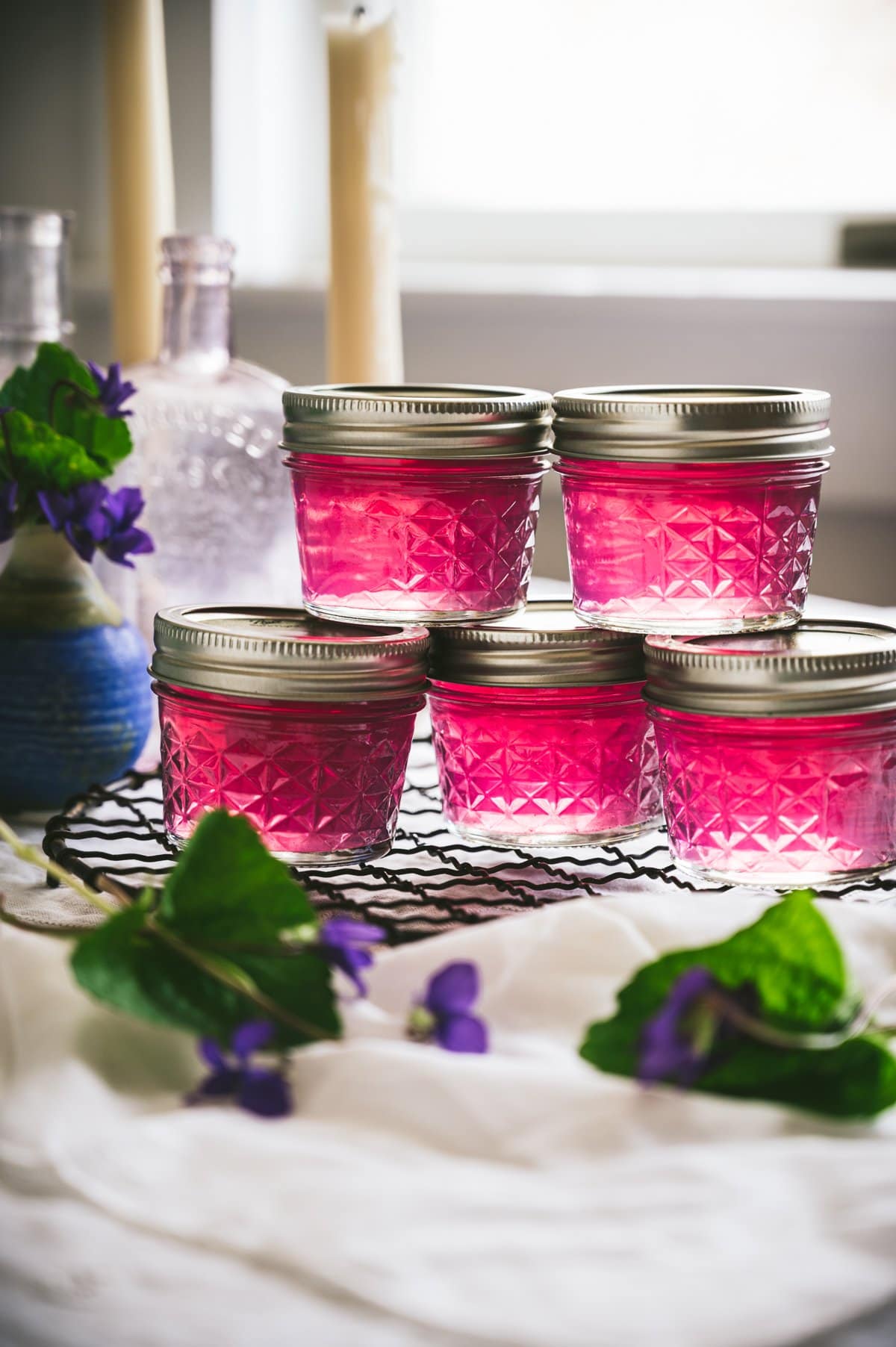 Stright on shot of 5 small quilted mason jars with metal screw top lids filled with purple/pink colored wild violet jelly stacked on top of each other.