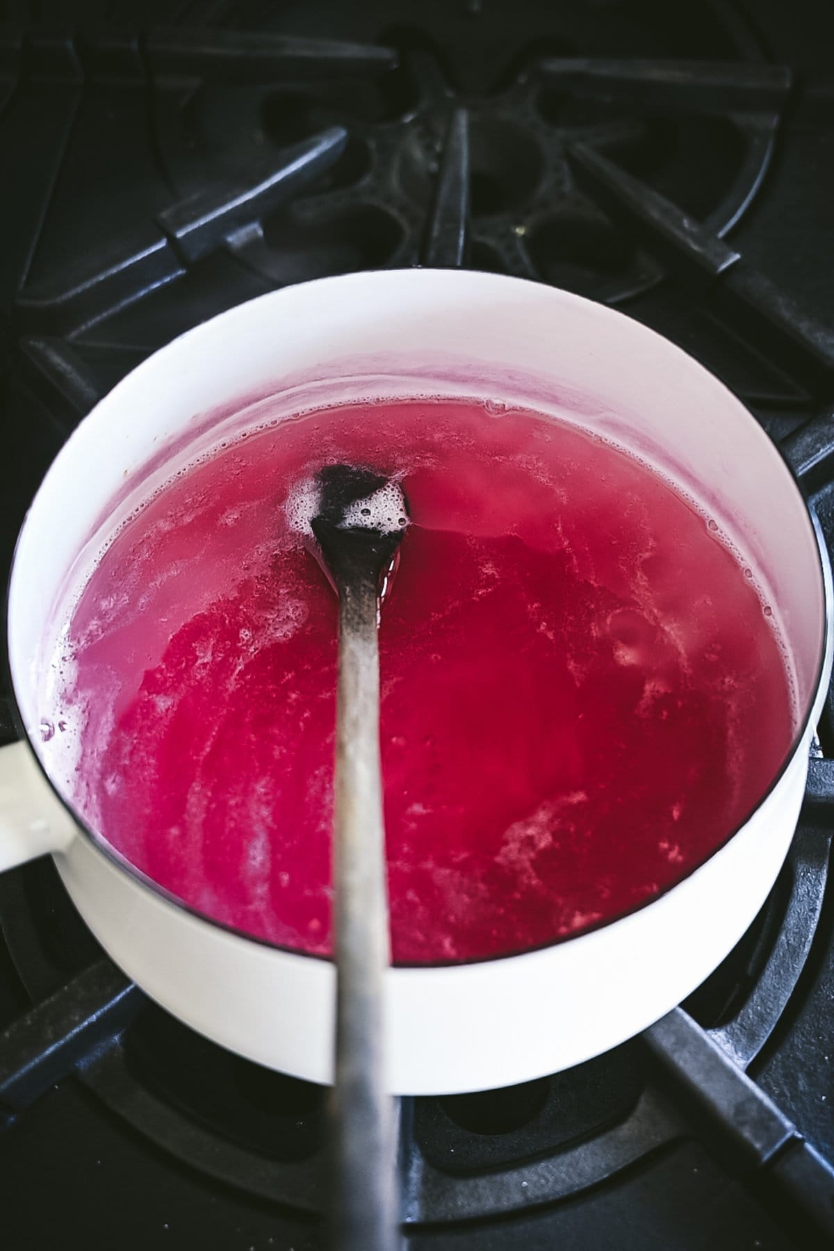 Cooked purple/pink coloered violet jelly in a white enamel saucepan on the stovetop with a dark walnut wood spoon in the pan.