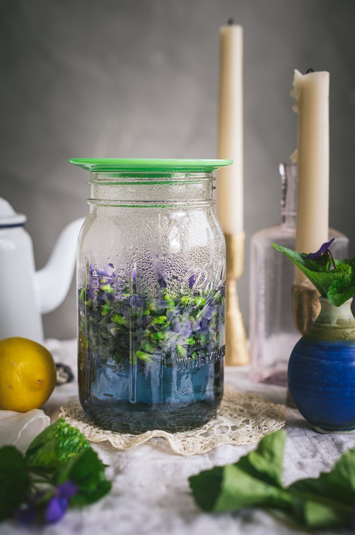 Straight on shot of violet flowers infusing into hot water in a glass quart mason jar with a small green glass plate covering the jar. The violet infusion has a blue color.