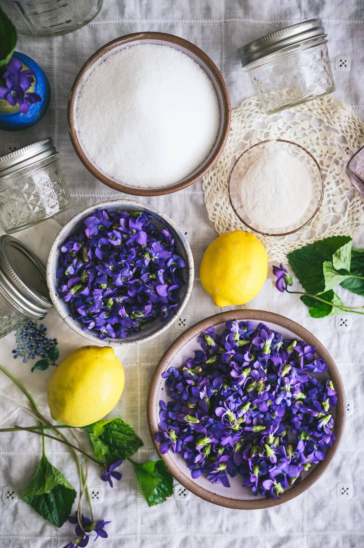 Top down shot of violet jelly ingredients measured out including fresh picked wild violet flowers, white sugar, powdered low-suagr pectin and lemons sitting on a white tablecloth surrounded by violet leaves, a small vase of violets and jelly jars.
