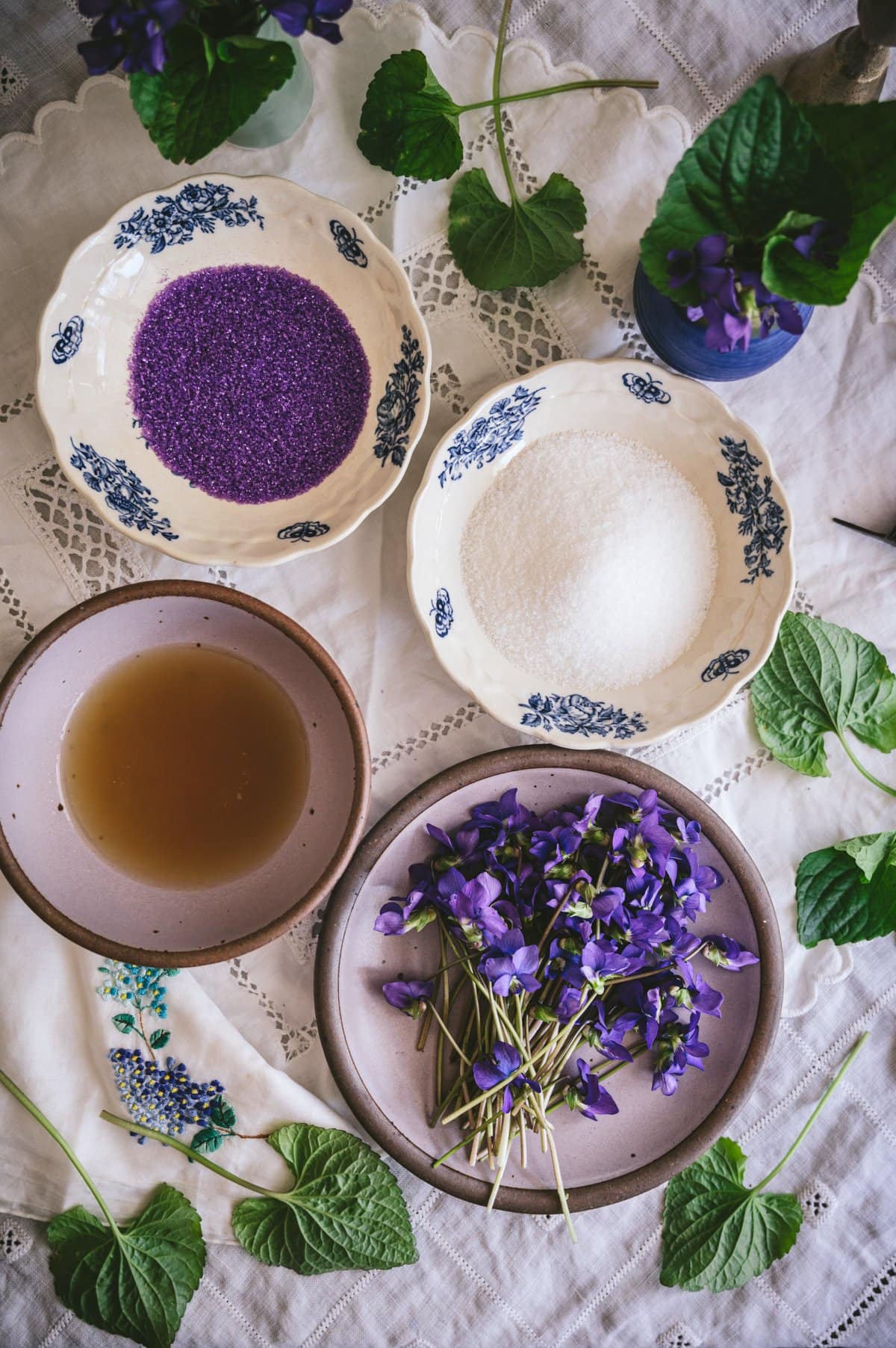 candied violets ingredients - wild blue violets on a purple plate, white sugar in a white dish, aquafaba in a purple bowl, and purple sanding sugar in a white dish.