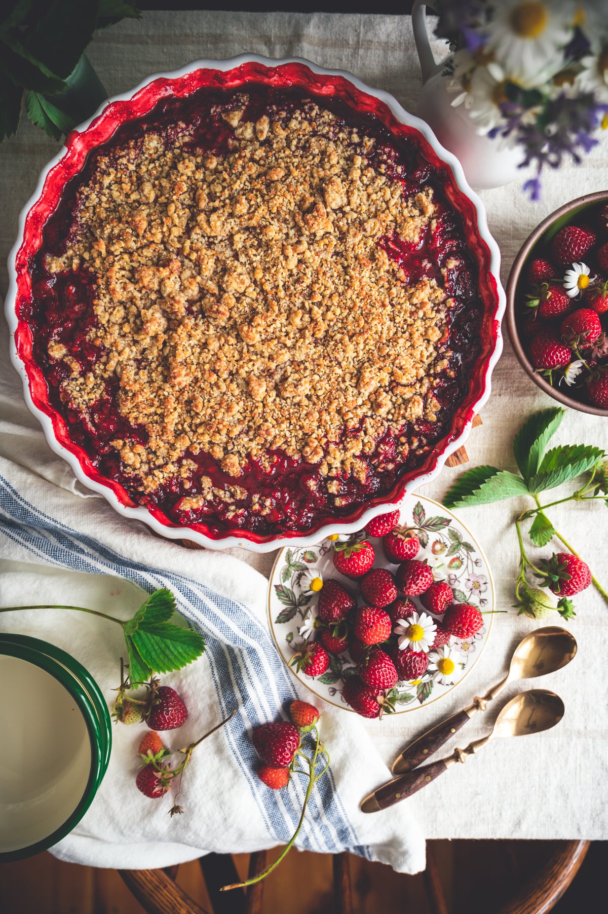 Cooked strawberry rhubarb crumble in a fluted tart pan on a table with fresh strawberries, bowls and spoons.