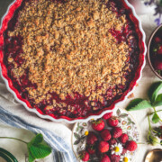 A top view of an easy strawberry rhubarb crumble in a scalloped baking dish, set on a table with a plate of fresh strawberries, green leaves, flowers, and a blue-striped cloth.