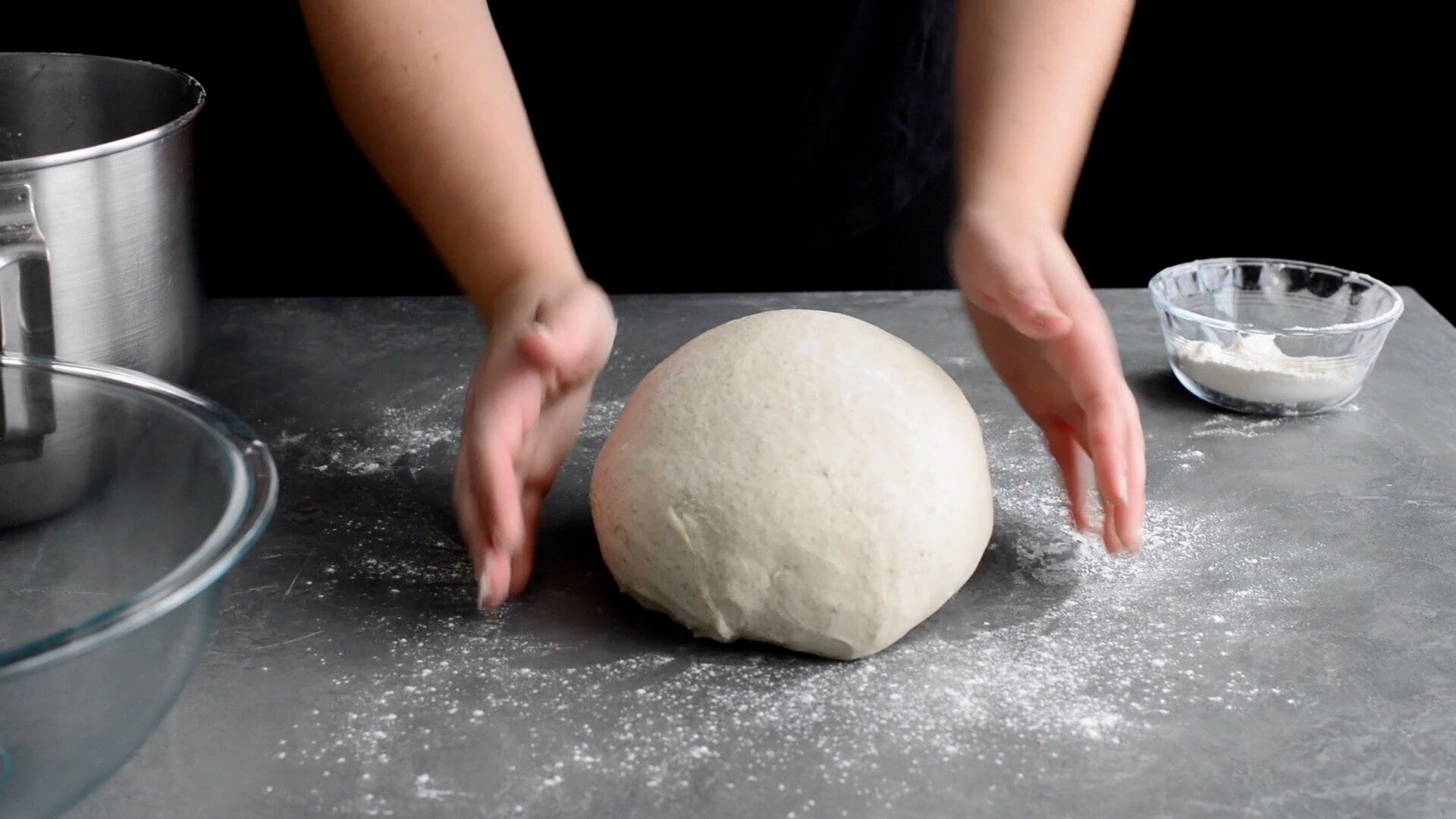 A person shapes a ball of dough on a floured countertop, preparing for a sourdough bagel recipe, with bowls of flour and utensils nearby.
