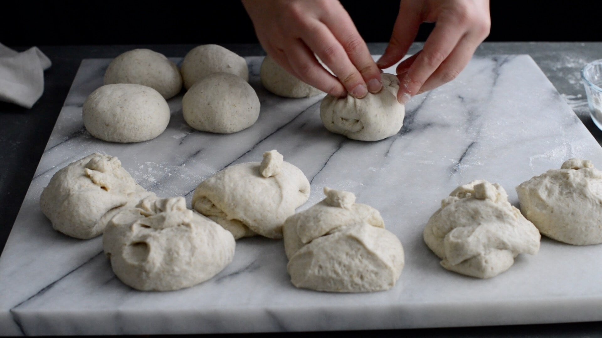 A person's hands shape pieces of dough into round balls on a floured marble surface, preparing for a sourdough bagel recipe, with several unshaped and shaped dough pieces visible.