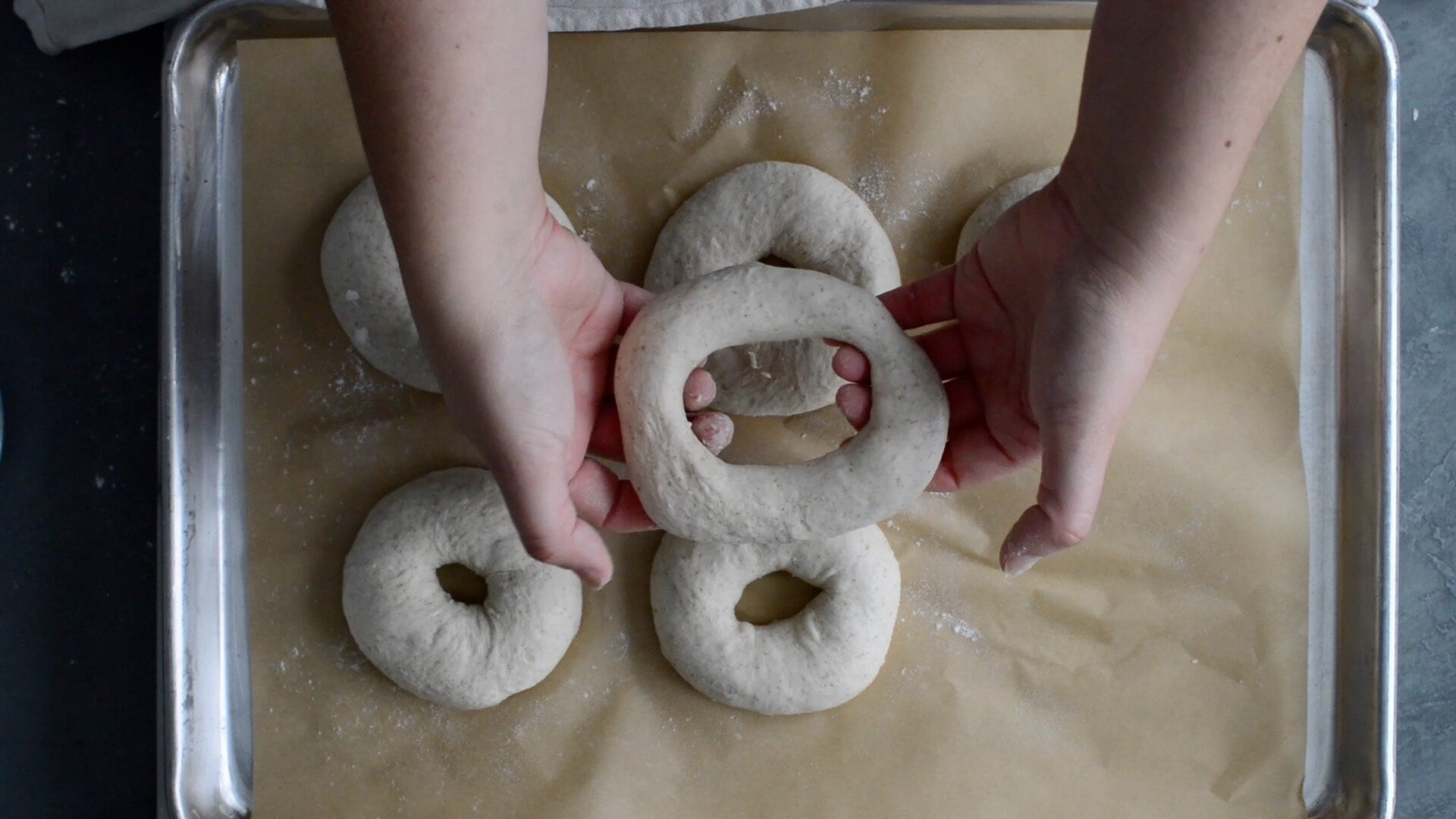 Hands holding a raw, ring-shaped sourdough bagel dough above a parchment-lined baking sheet with several other uncooked bagels, ready for baking—perfect for your next sourdough bagel recipe adventure.