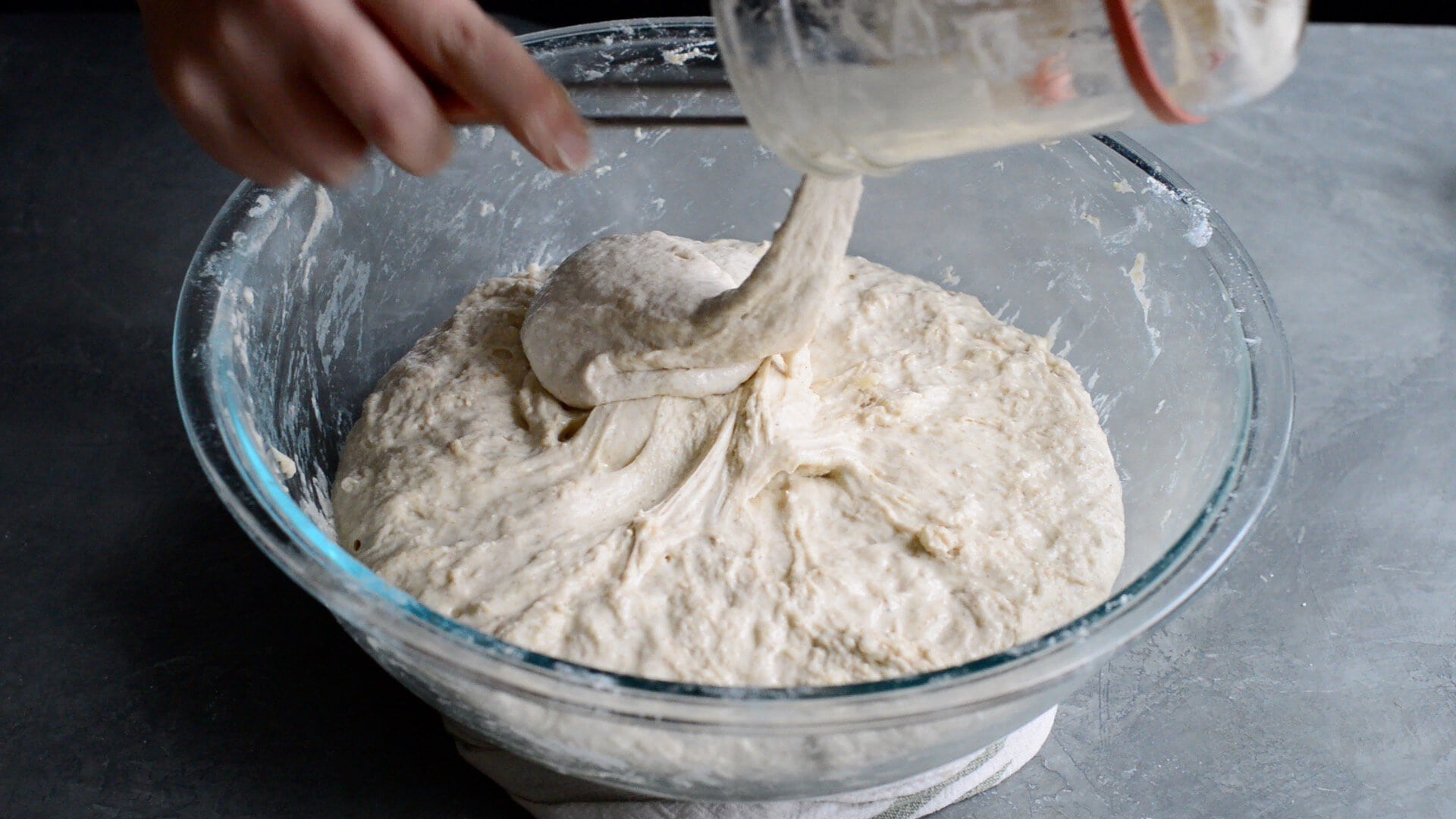 A hand is pouring a sticky sourdough starter from a glass jar into a large bowl of autolyzed dough, an essential step to an artisan sourdough bread recipe, all set on a gray surface.