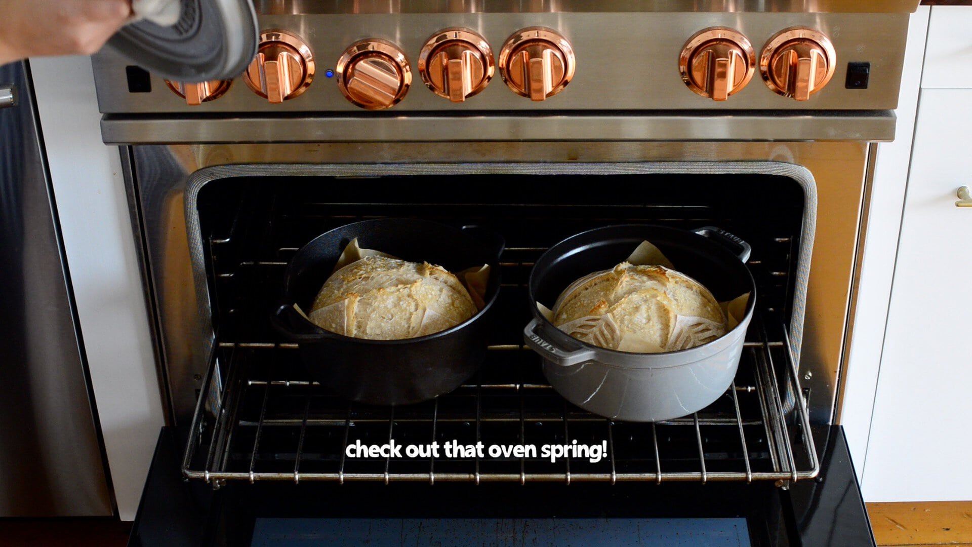Two round loaves from our artisan sourdough bread recipe bake in Dutch ovens in an oven range with copper knobs. Text on the image reads, "check out that oven spring!.