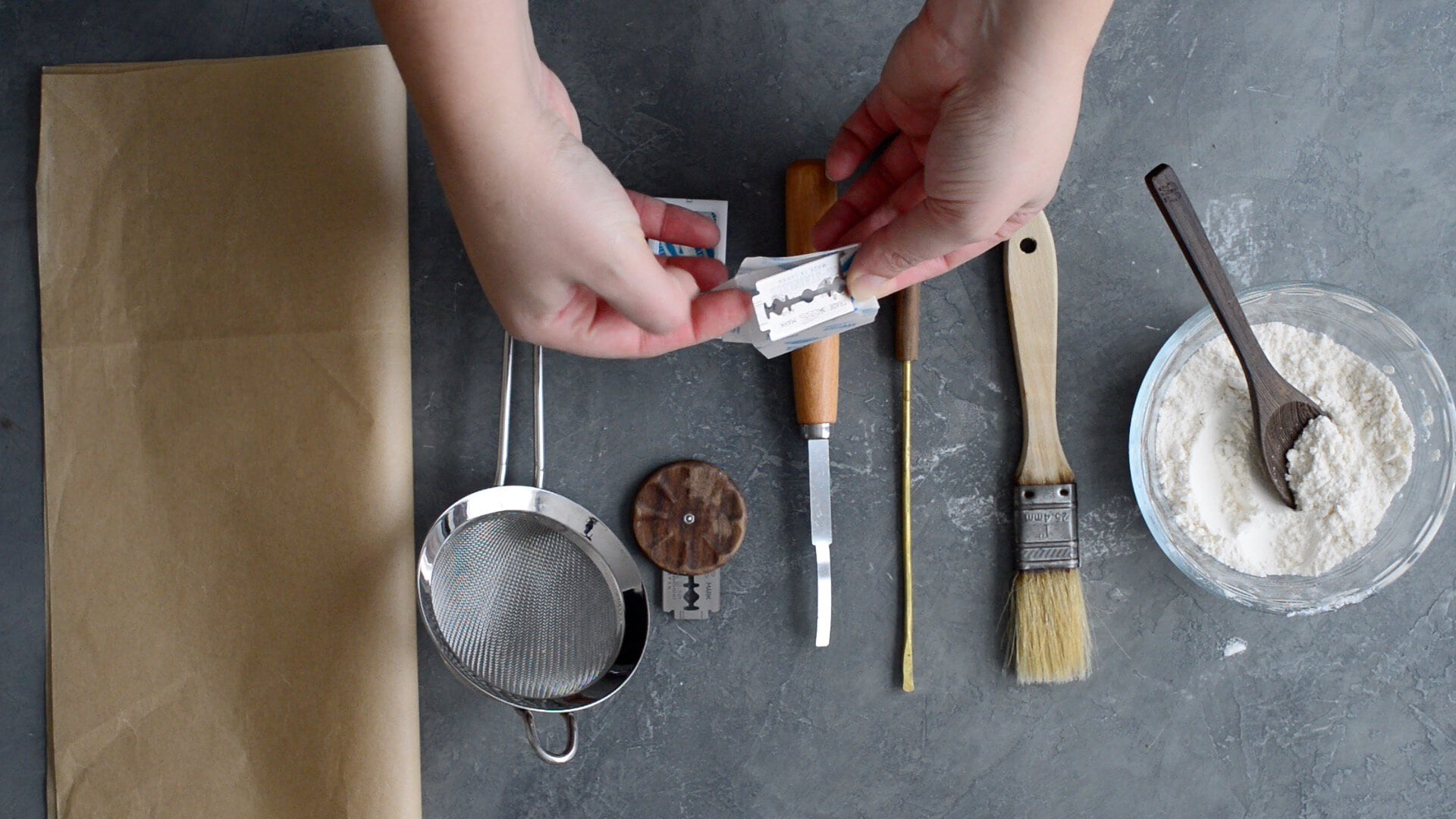 Hands unwrapping a razor blade above a table set for baking—a sourdough bread recipe in progress with parchment paper, strainer, wooden tool, knife, brushes, skewer, and a bowl of flour on a gray surface.
