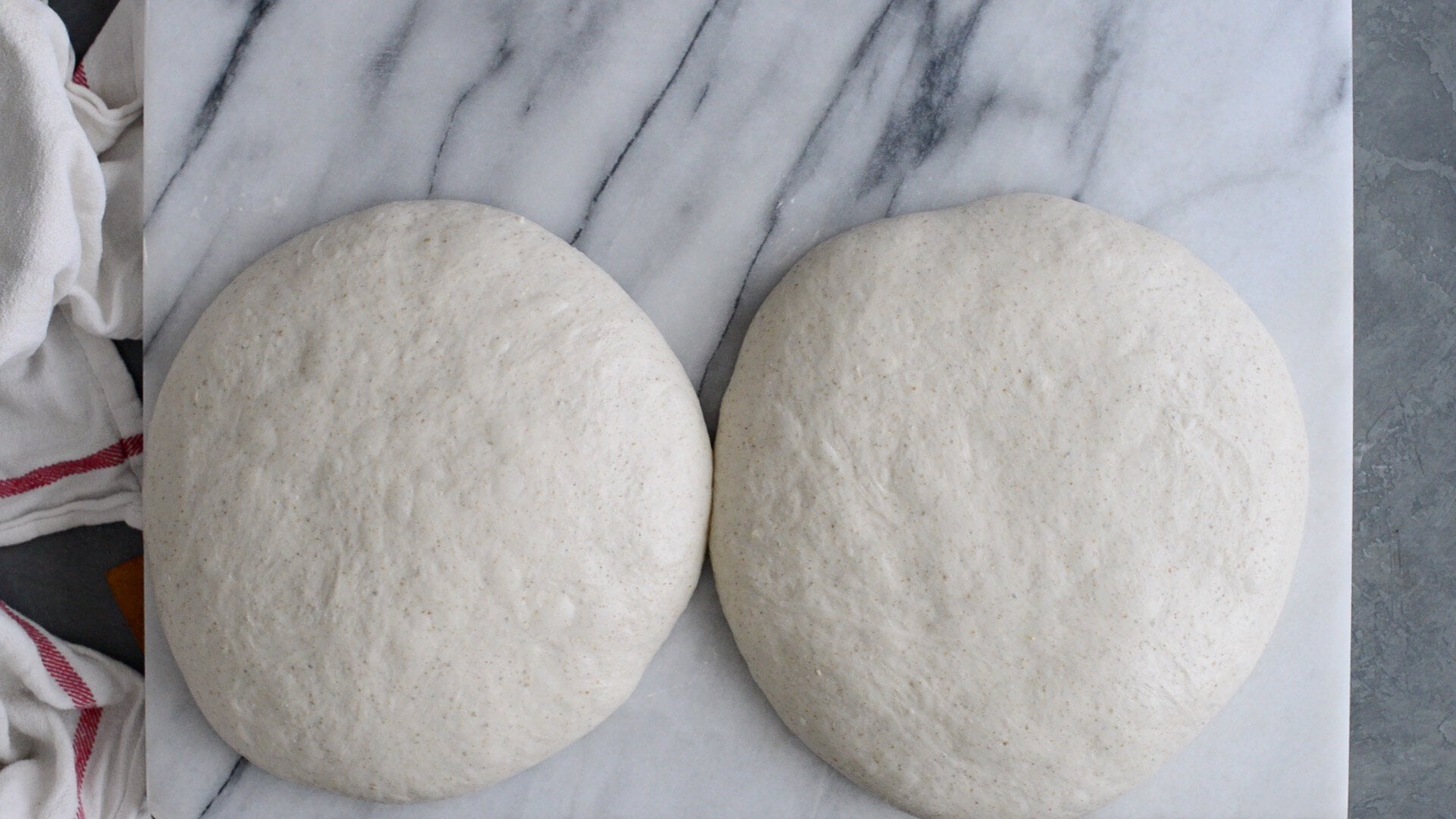 Two round balls of bench rested artisan sourdough dough, made from a sourdough bread recipe, rest on a white marble surface. A white cloth with red stripes is visible on the left edge of the image.