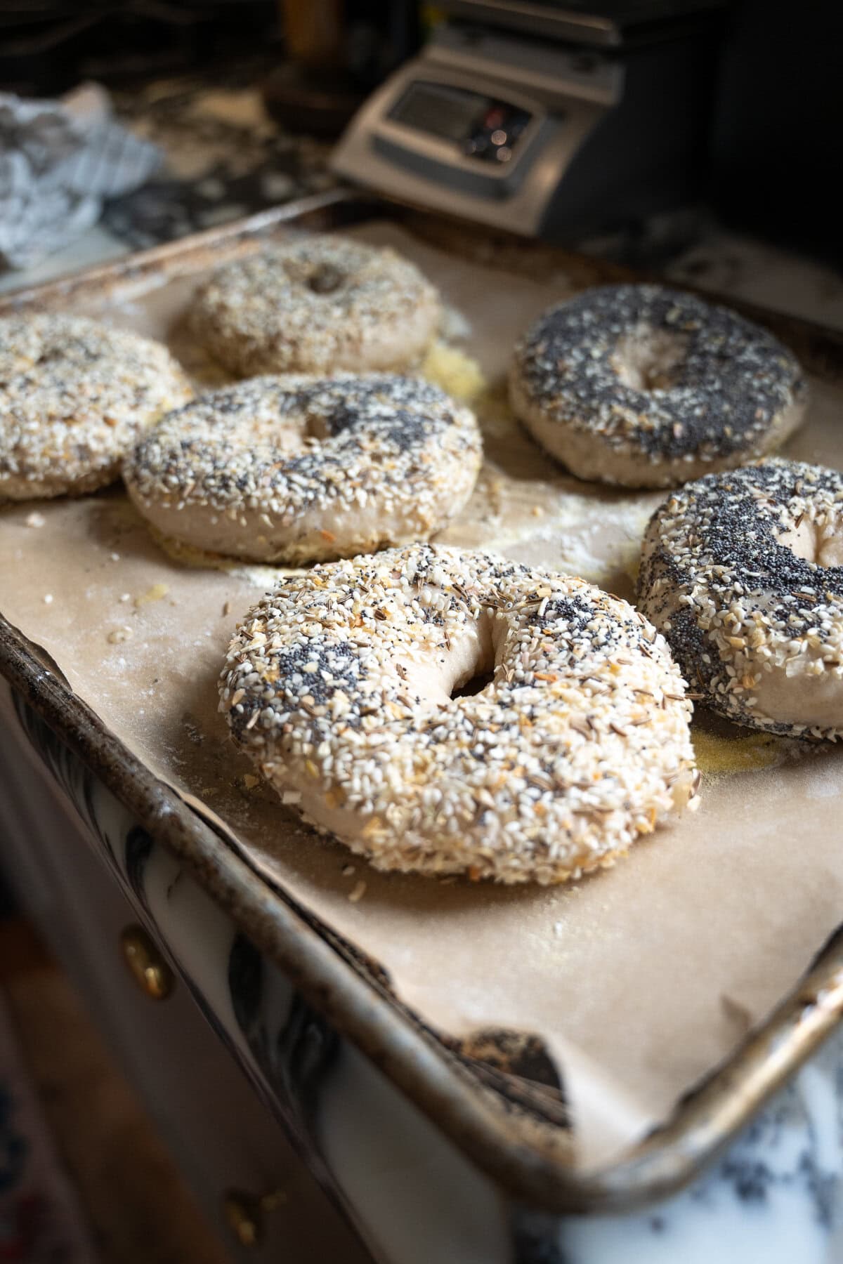 Six unbaked sourdough bagels covered in seeds are arranged on a parchment-lined baking sheet, ready to be baked. A digital scale and other kitchen items sit in the background, completing this sourdough bagel recipe scene.