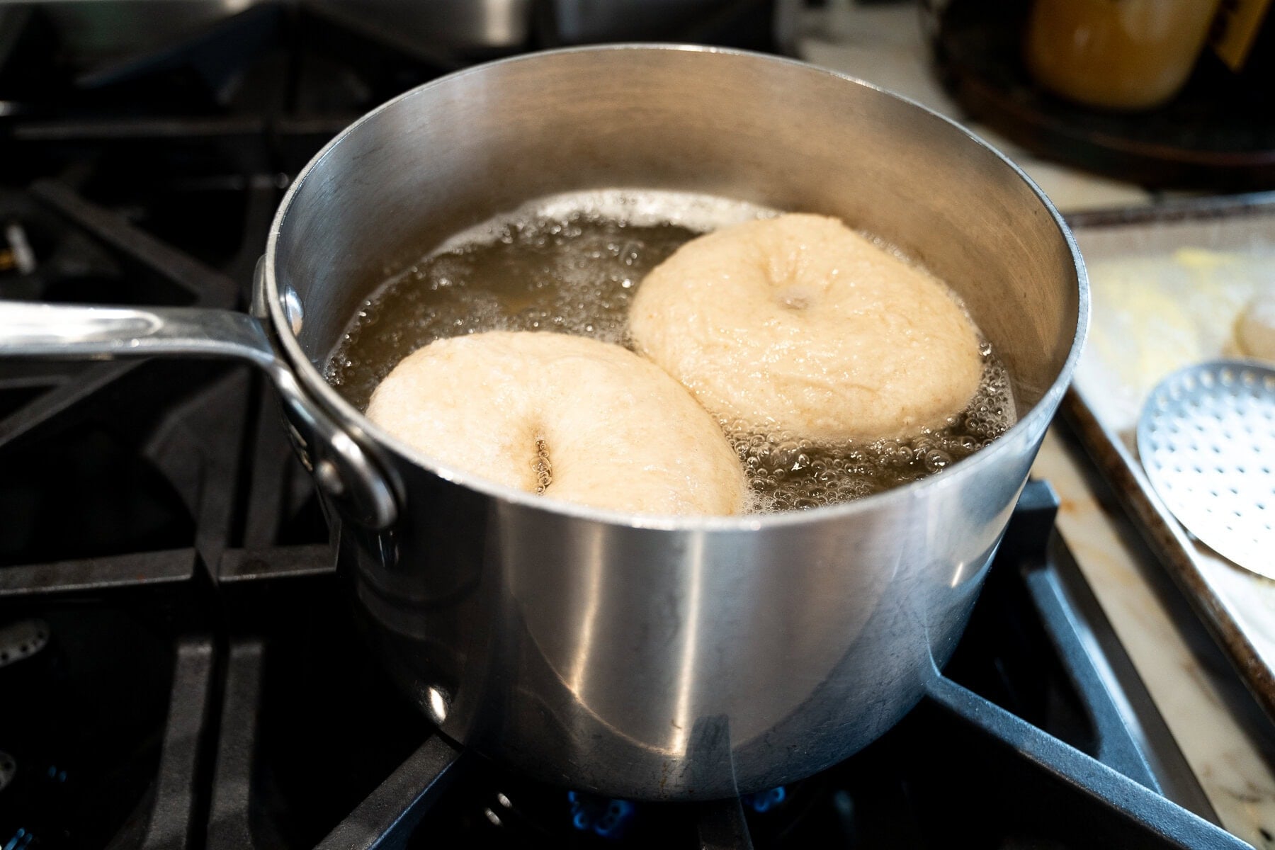 Two raw bagels are boiling in a pot of water on a stovetop as part of the sourdough bagel recipe process. The pot sits on a gas burner, with a slotted spatula visible nearby.
