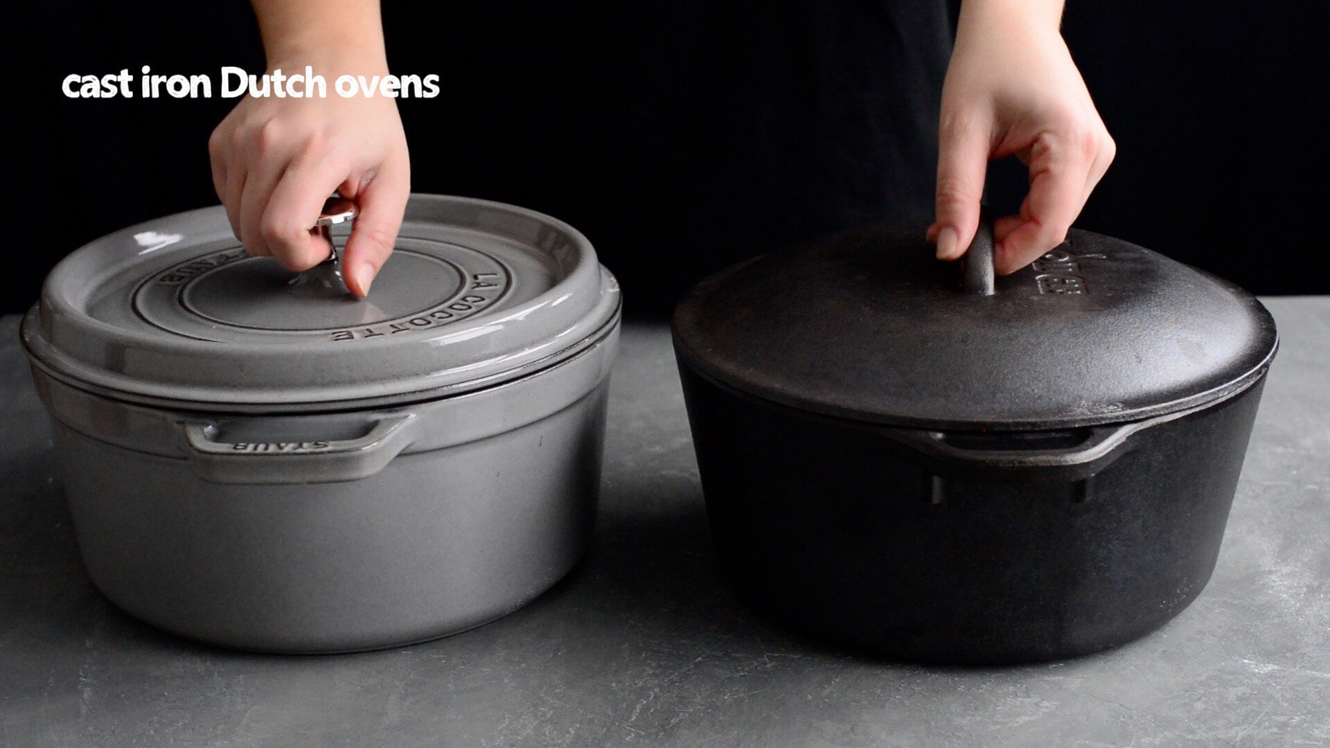 Two hands lift lids off two cast iron Dutch ovens, perfect for baking an artisan sourdough bread recipe, one gray and one black, placed side by side on a gray surface with the text "cast iron Dutch ovens" in the top left corner.