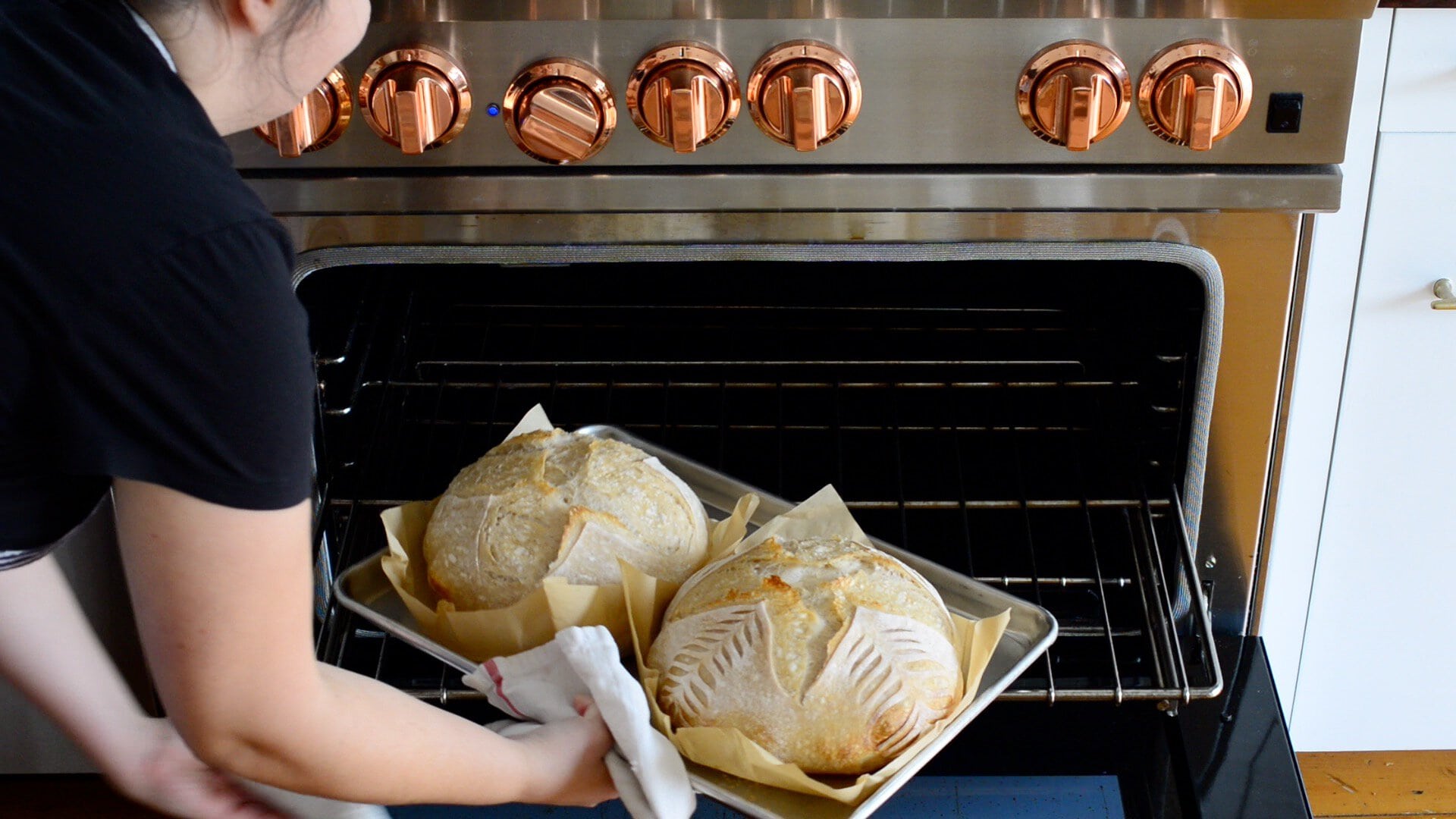 A person places a baking tray with two round loaves, prepared using an artisan sourdough bread recipe, into an oven with shiny copper knobs. The loaves rest on parchment paper, looking partially baked and ready to finish baking uncovered.