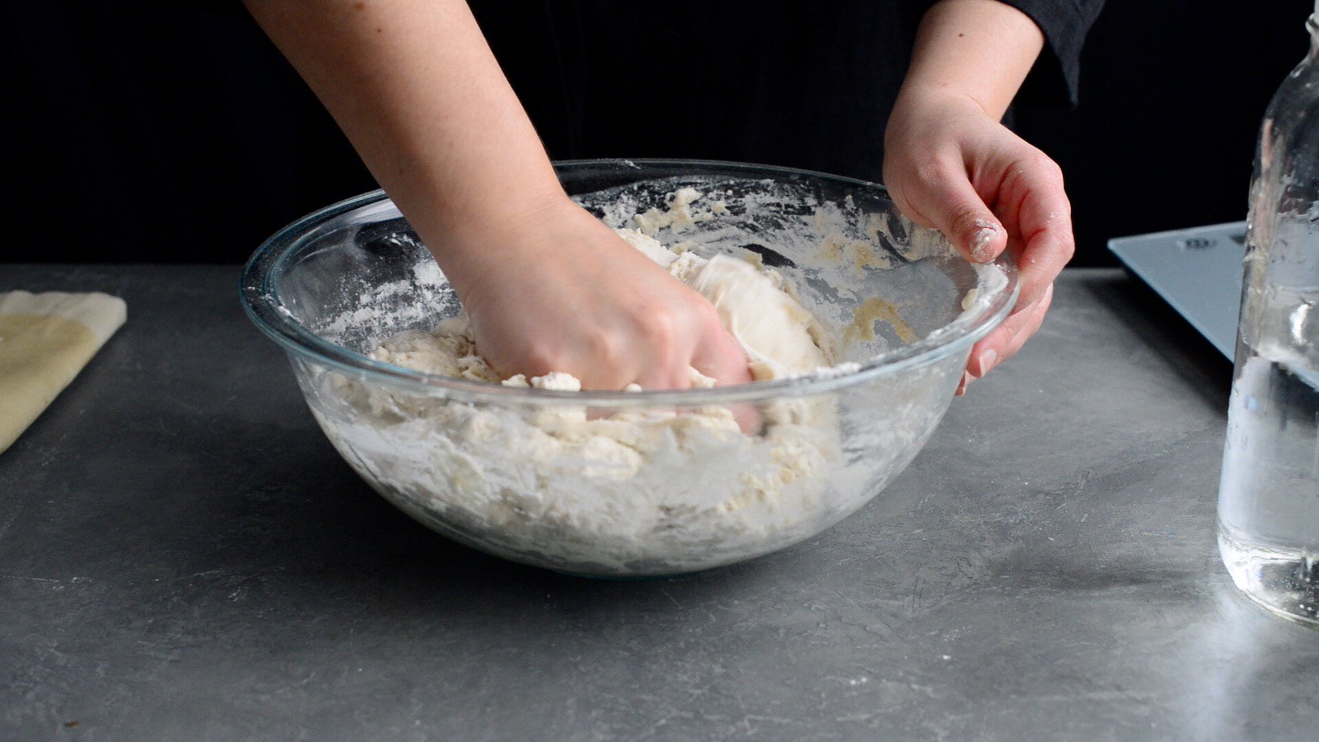 A person kneads dough in a glass bowl on a gray countertop, following an artisan sourdough bread recipe, with a bottle of water and a laptop nearby.
