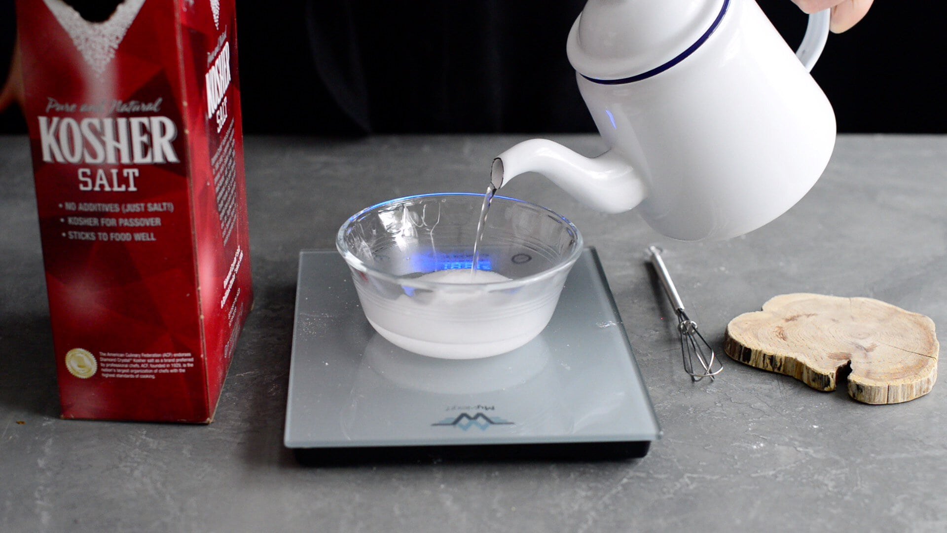 A person pours hot water from a white kettle into a glass bowl with salt on a digital kitchen scale—a steps in an artisan sourdough bread recipe. Kosher salt, a mini whisk, and a wooden coaster are nearby on the gray countertop.