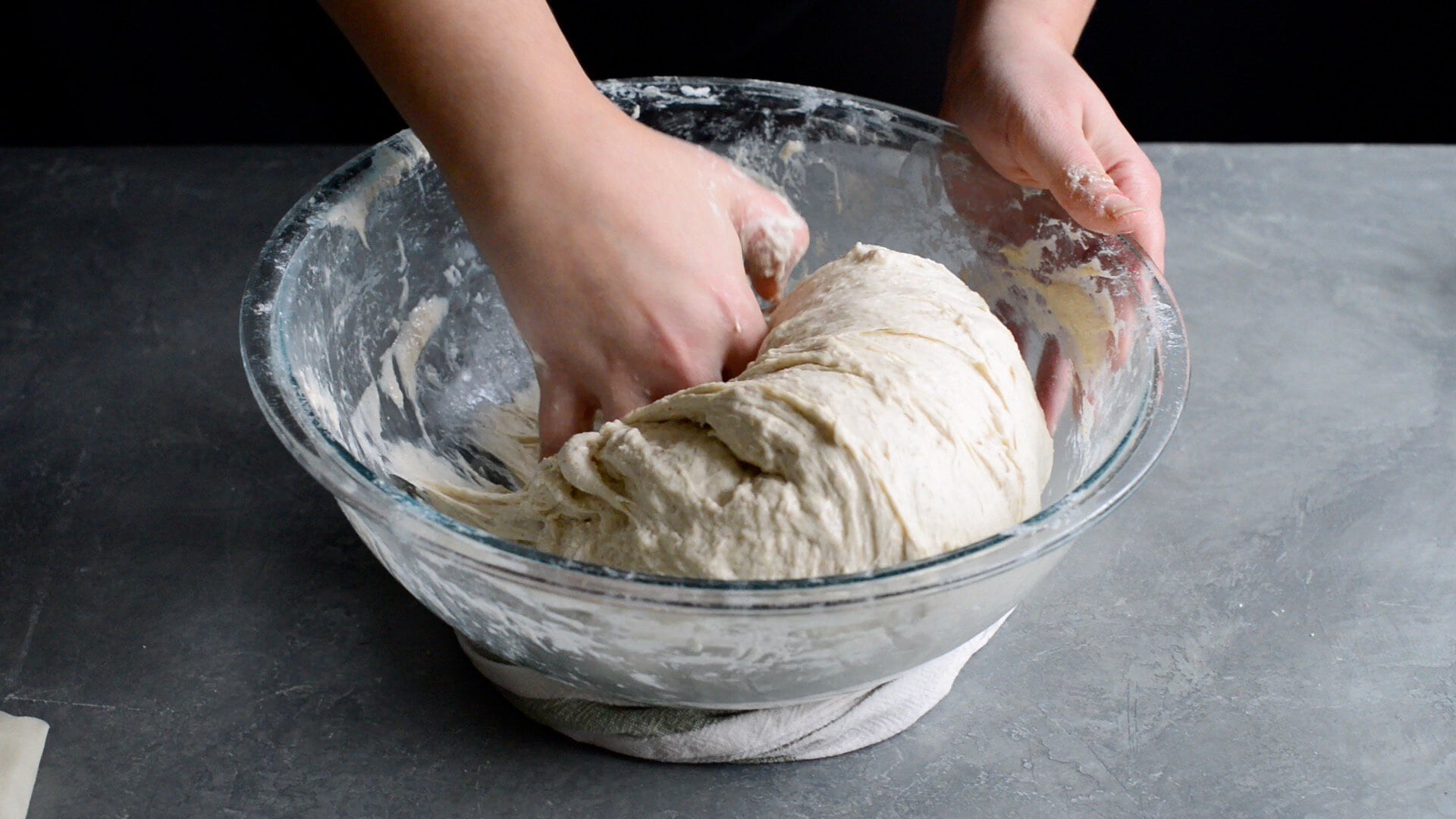 A person kneads salt and water into dough in a large glass bowl on a gray countertop, following an artisan sourdough bread recipe, with flour scattered on their hands and the bowl’s rim.