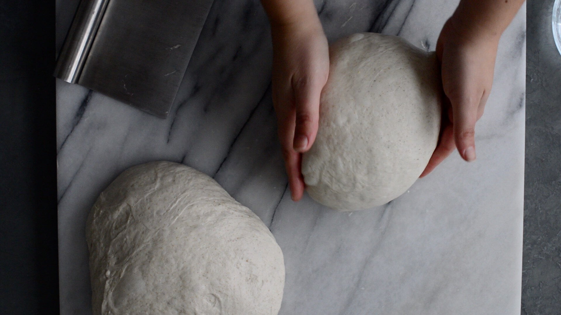 A person preshaping a ball of artisan sourdough bread dough on a marble surface, following a sourdough bread recipe, with another dough ball and a metal dough scraper nearby.