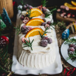 Cranberry icebox cake covered in whipped cream and decorated with sugared cranberries, orange slices, sliced cranberries and rosemary sprigs on a white rectangular platter.