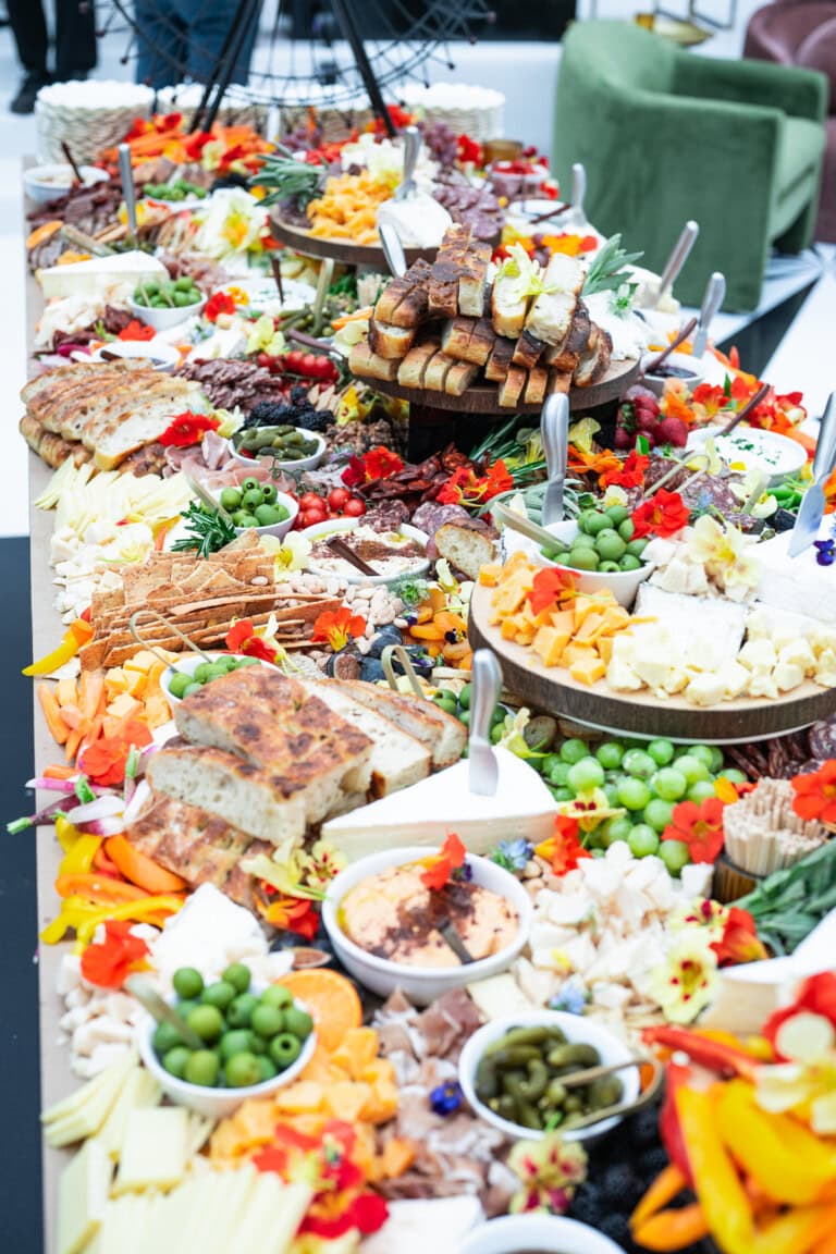 A stunning grazing table brimming with a diverse assortment of colorful foods—cheeses, meats, fruits, vegetables, and baked goods—perfect for a Nantucket wedding celebration. In the background, plush green and pink seating complements the geometric floor patterns elegantly.