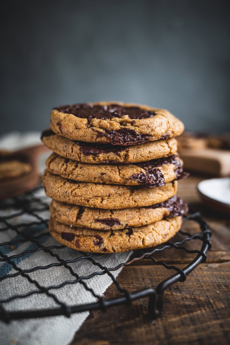 A stack of six whole wheat chocolate chip cookies sits on a black wire cooling rack, with melted chocolate visible in the cookies. The background is blurred, featuring a rustic wooden surface and fabric napkin.