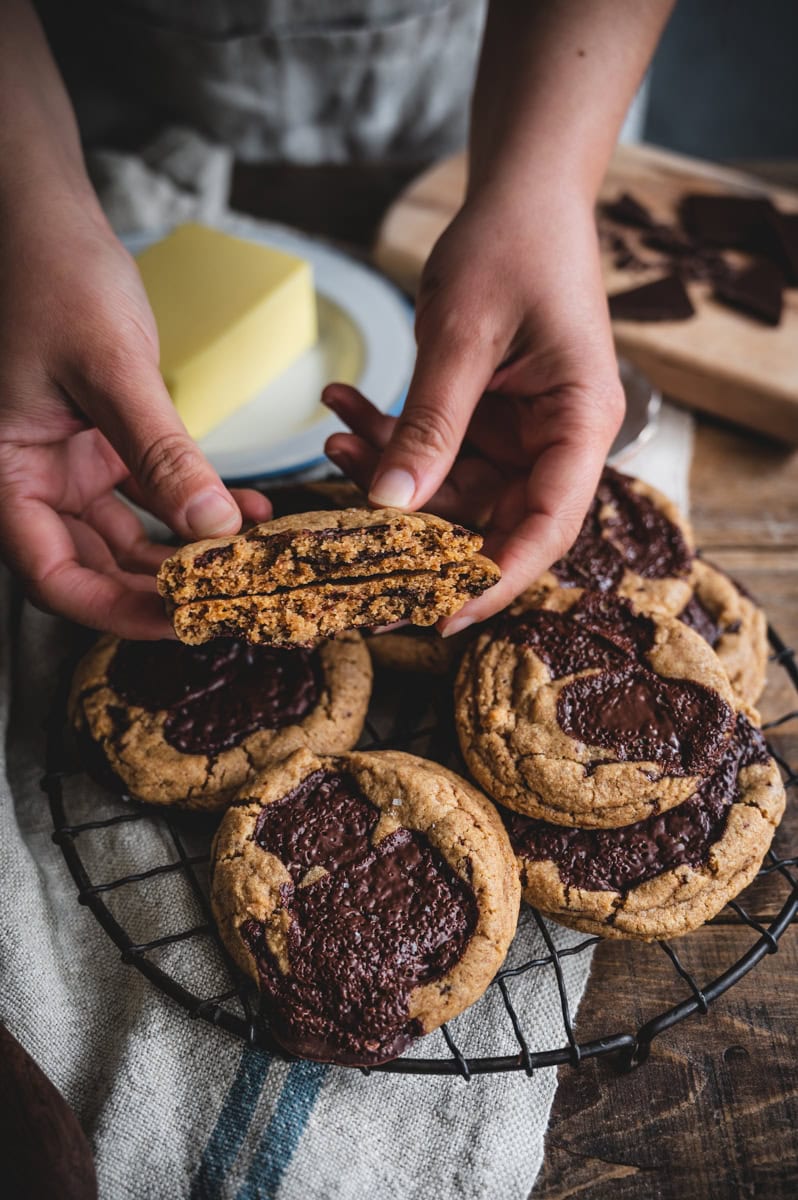 A person holds a split whole wheat chocolate chip cookie above a cooling rack filled with large cookies topped with melted chocolate. A plate with a block of butter and a cutting board are in the background.