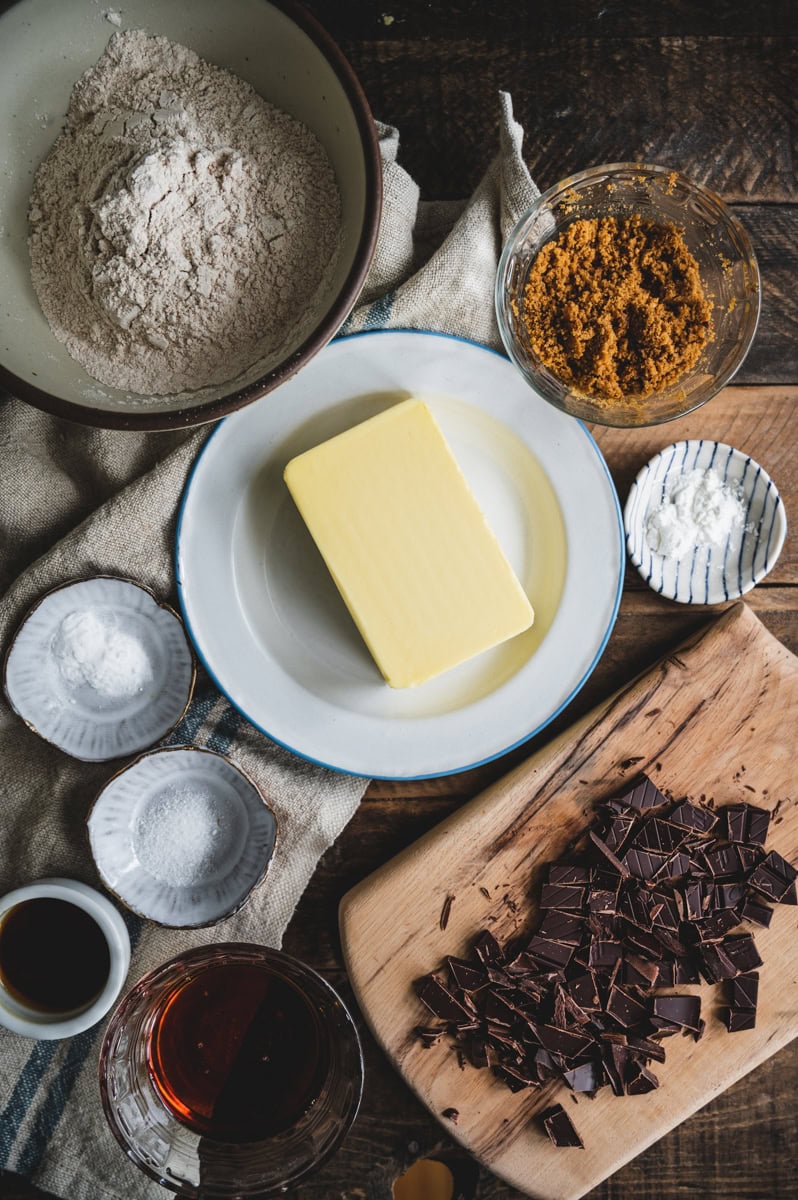 A flat lay of baking ingredients for whole wheat chocolate chip cookies on a rustic table, including a stick of butter, flour, brown sugar, chopped chocolate, baking powder, salt, vanilla extract, and liquid sweetener.