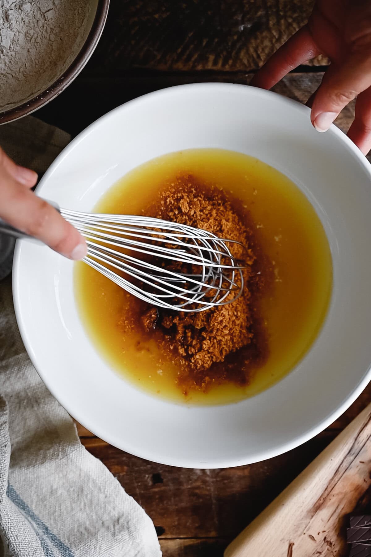 A person uses a whisk to mix brown sugar into melted butter in a white bowl on a wooden surface, preparing ingredients for whole wheat chocolate chip cookies.