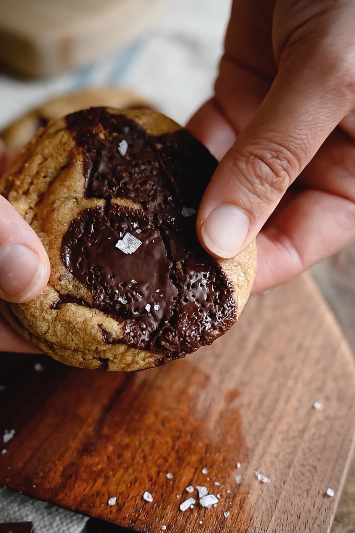 A close-up of hands holding a thick whole wheat chocolate chip cookie topped with sea salt flakes, over a wooden surface.