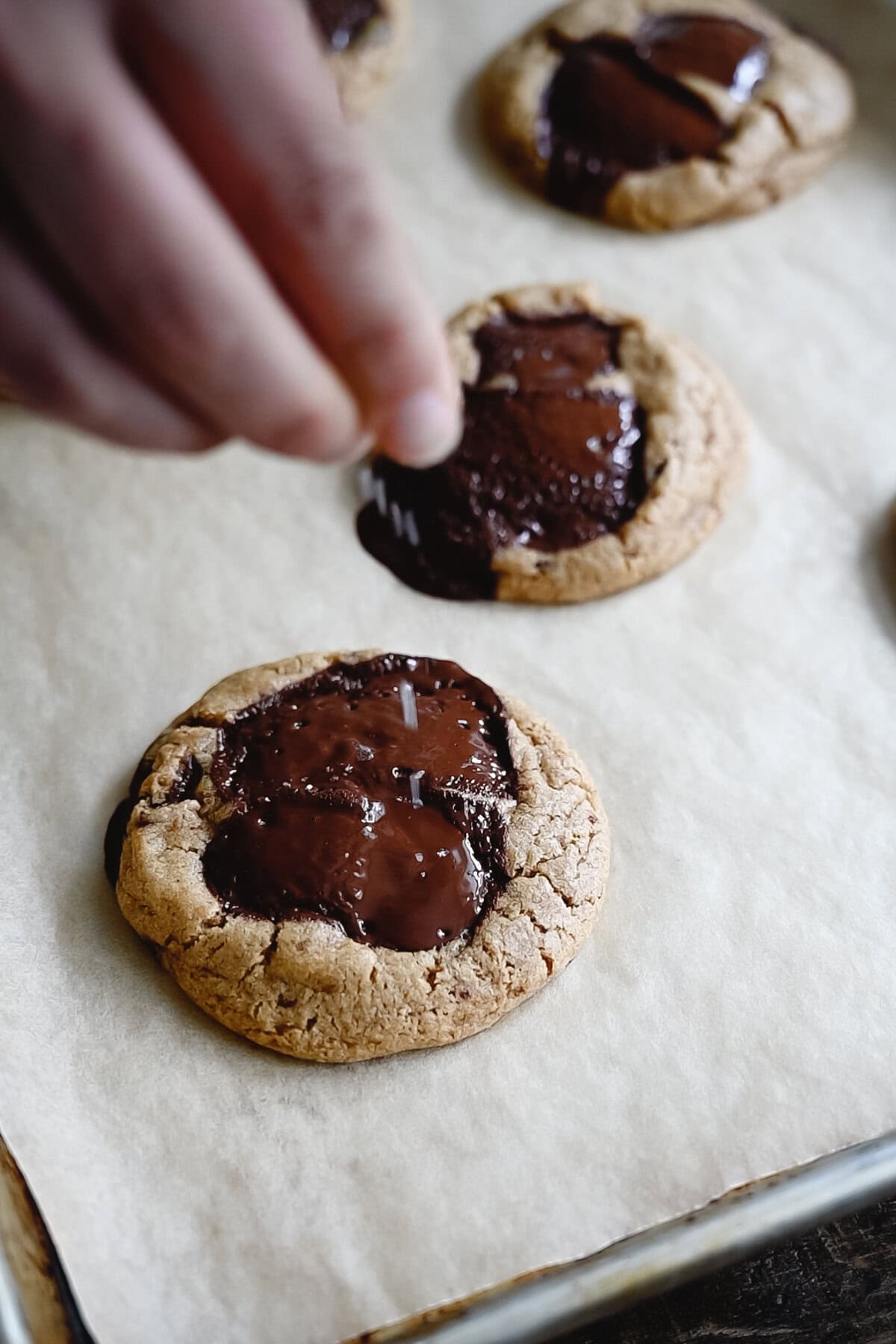 A hand sprinkles flaky salt onto a freshly baked whole wheat chocolate chip cookie topped with melted chocolate, resting on parchment paper alongside other cookies.