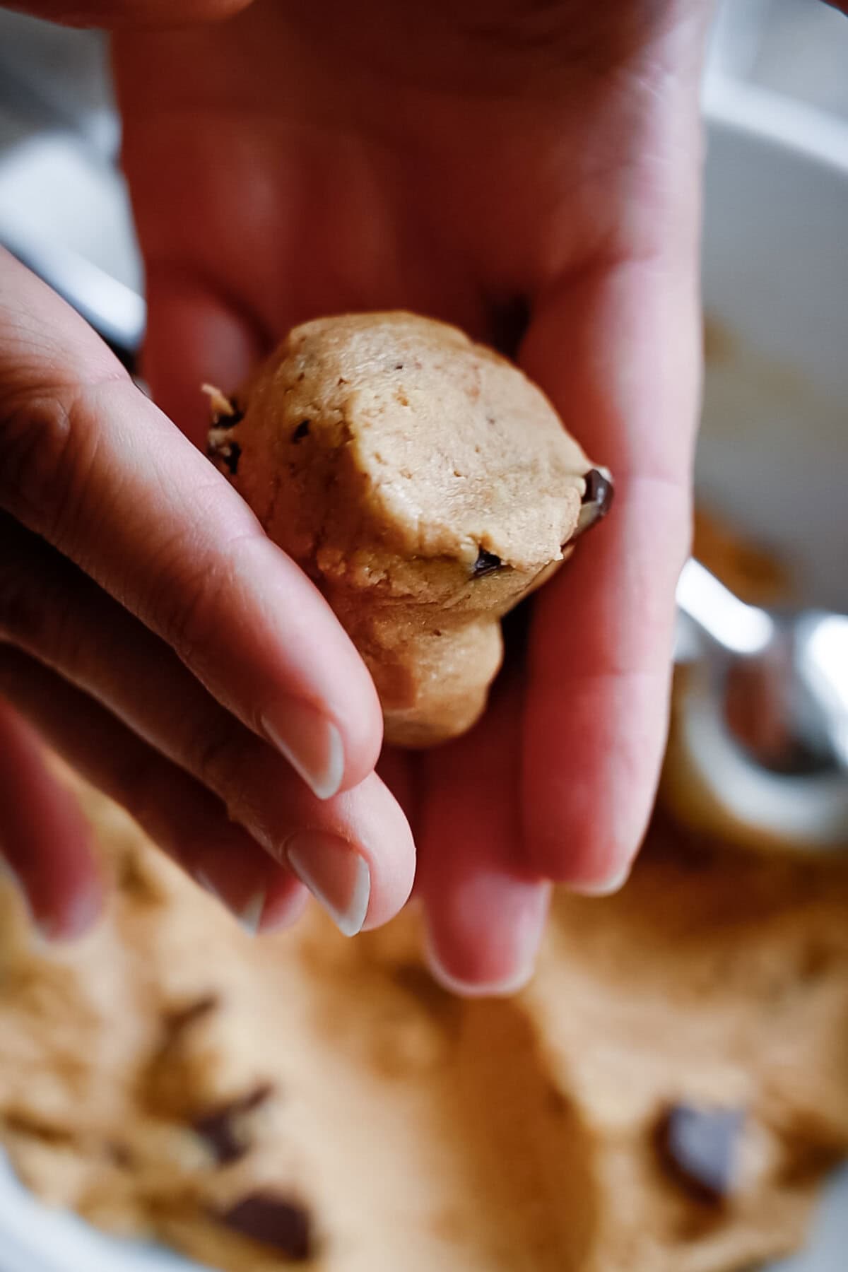 A close-up of hands holding a ball of whole wheat chocolate chip cookie dough, with more dough and a metal spoon blurred in the background.