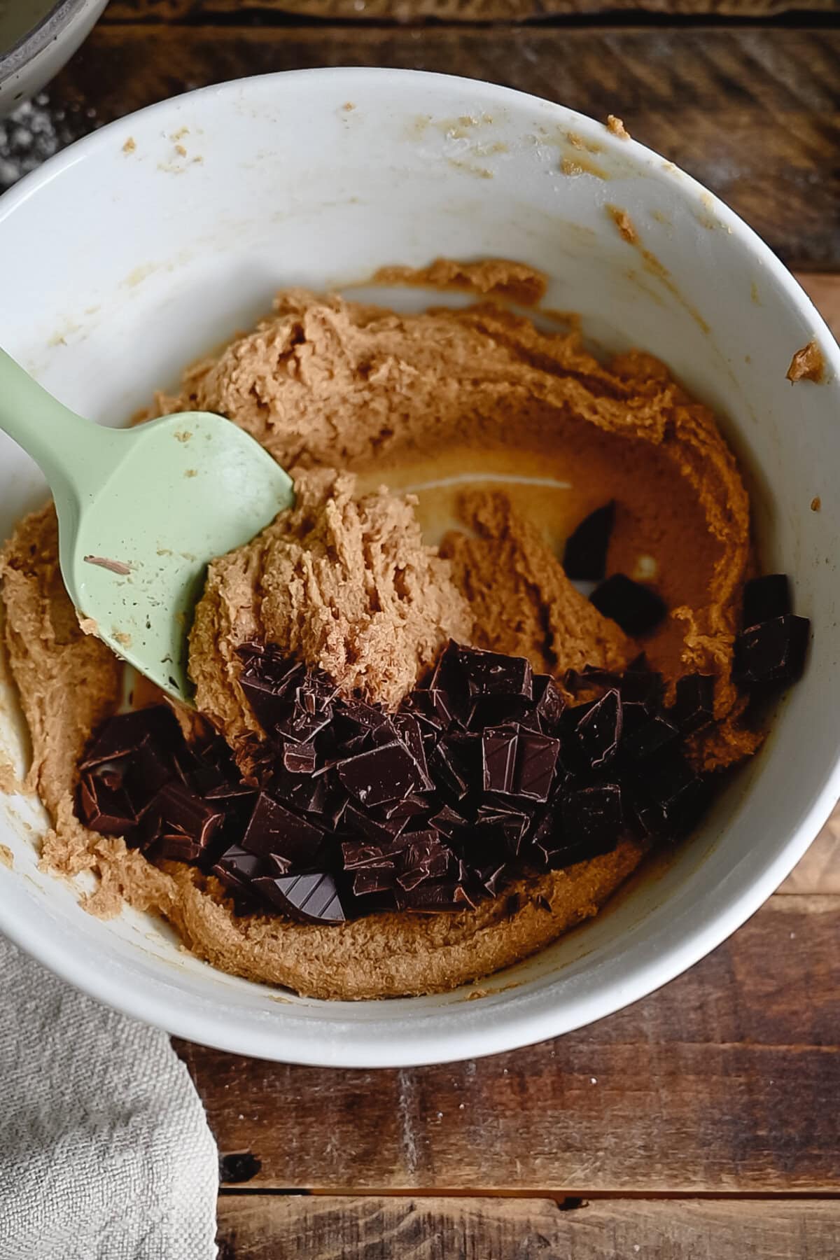 A white mixing bowl filled with whole wheat chocolate chip cookie dough and dark chocolate chunks sits on a wooden surface. A green spatula rests inside the bowl, ready to mix the ingredients together.