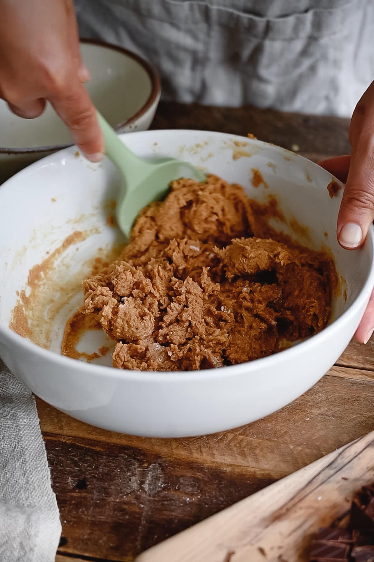 A person mixes whole wheat cookie dough with a green spatula in a white bowl on a wooden table, preparing ingredients for baking.