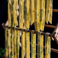 Wide strips of yellow pappardelle pasta with green herbs pressed inside hang evenly across wooden rods, ready to dry. The dark background lets this fresh pappardelle pasta recipe stand out beautifully.