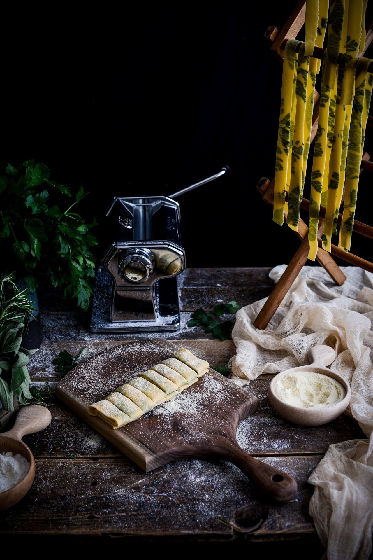 A rustic kitchen scene with a pasta machine, fresh herb pasta drying on a rack, rolled ravioli on a floured wooden board, bowls of flour and cheese, and fresh herbs on a wooden table.