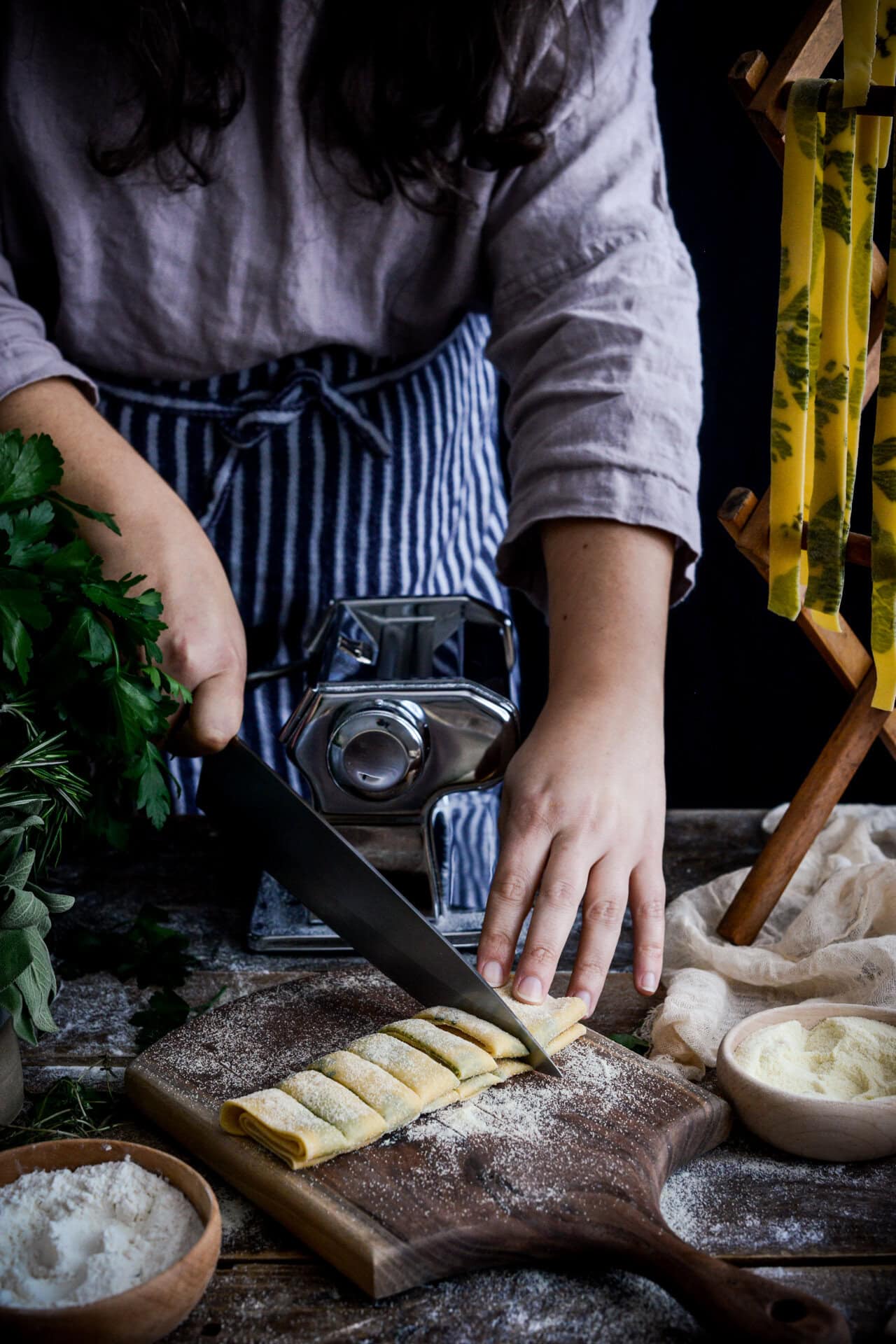 A person in a striped apron slices rolled, fresh pasta dough on a floured board, with a pasta machine, herbs, and drying pasta visible nearby.